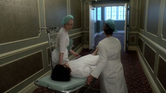  GaGa is lying on a gurney being wheeled through a hallway por two nurses wearing mint Parisian berets.
